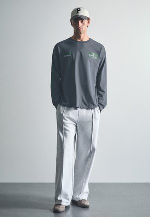 Jeune homme debout, les mains dans les poches, portant un t-shirt à manches longues sombre avec le logo Toyota, un pantalon blanc, des chaussures grises et une casquette blanche avec la lettre P.