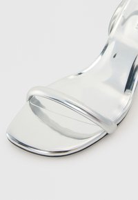 Ασημί σανδάλι με τακούνι και λεία, ανακλαστική επιφάνεια, με δύο λεπτές λωρίδες και σχέδιο τετράγωνης μύτης. Μαύρη σόλα τονίζει την εμφάνιση.