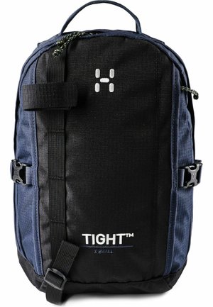 Mochila compacta en negro y azul oscuro con asa superior, hebillas laterales, correas frontales y logo "TIGHT™ X SMALL" en el panel frontal.
