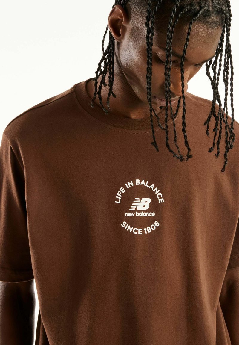 Young man with braided hair wearing brown New Balance T-shirt with "Life in Balance Since 1906" logo, looking down against a plain background.