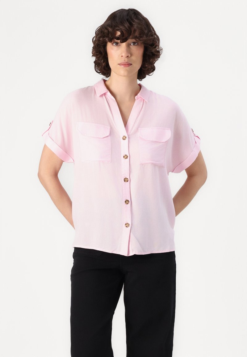 Chemise courte à manches courtes de couleur rose clair avec un col, deux poches avant et des boutons en bois. Tissu doux et léger avec une coupe décontractée.