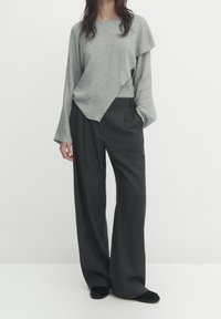 Grijze, textuurgegeven, lange mouw top met een asymmetrische zoom, gecombineerd met high-waisted, wijde, donkergrijze broek met subtiele strepen. Zwarte platte schoenen.