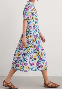 Bloemenpatroon, korte mouwen, jurk tot op de knie van lichte stof, met paarse, gele en blauwe accenten; gecombineerd met bruine sandalen met bandjes.