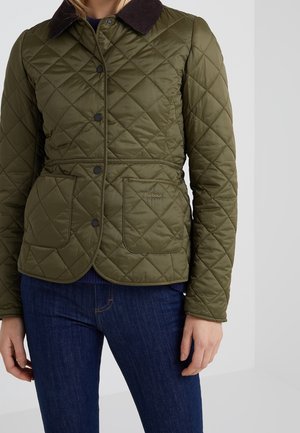 Personne portant une veste matelassée verte avec un col noir, des boutons pression et des poches avant, associée à un jean bleu foncé.