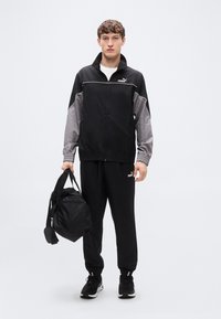 Μαύρο και γκρι αντιανεμικό με λογότυπο, ταιριαστό μαύρο παντελόνι φόρμας και μαύρα αθλητικά παπούτσια. Το μοντέλο κρατά μια μαύρη τσάντα duffel.