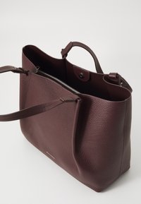 Sac fourre-tout en cuir de couleur bourgogne foncé avec surface texturée, ouverture en haut, deux poignées courtes et une poche intérieure zippée. Détail de la marque subtil au bas.