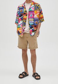 Kurzärmliges, mehrfarbiges Hemd mit geometrischen Mustern, kombiniert mit khakifarbenen Shorts und schwarzen Schlappen.