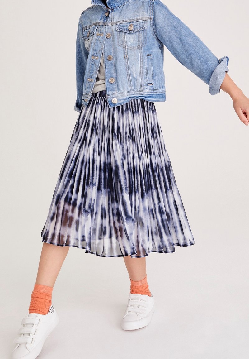 Fille portant une jupe plissée noir et blanc tie-dye, une veste en jean bleu clair, des chaussettes orange et des baskets blanches, debout devant un fond uni.