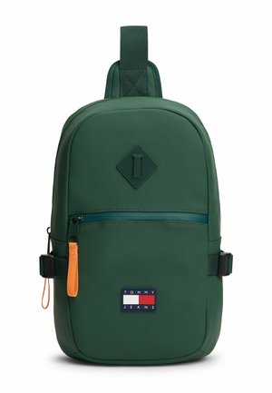 Dark green Tommy Jeans sling backpack with front zip pocket, orange zipper pull, adjustable side straps, and diamond-shaped detail.