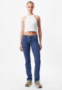 Jeans de mezclilla azul con un diseño ajustado y un ligero estiramiento, combinados con una blusa sin mangas corta blanca y zapatillas beige.
