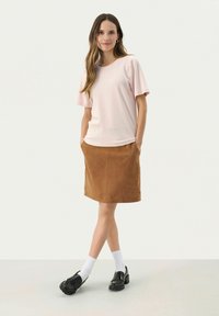Top rosa chiaro a maniche corte abbinato a una gonna in suede marrone. La modella indossa mocassini neri e calzini bianchi. Design semplice, texture morbide.