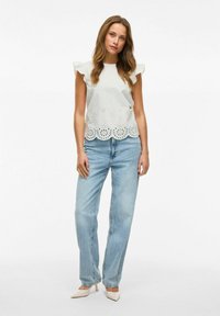 Lys grå top med flæseærmer og broderet bølgekant; parret med lys blå lige ben jeans og beige spidse hæle.