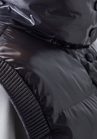 Schwarze Steppweste mit glänzender, gepolsterter Textur, gerippten Schulterträgern und Druckknopfverschluss, der ein elegantes, modernes Design unterstreicht.