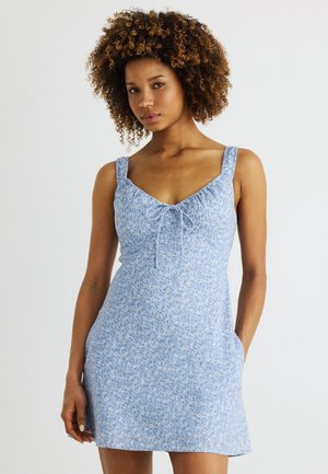 Γυναίκα με σγουρά μαλλιά που φοράει ένα αμάνικο ανοιχτό γαλάζιο φόρεμα με λουλούδια και λεπτομέρεια δέσιμου στο λαιμό, όρθια μπροστά σε έναν απλό φόντο.