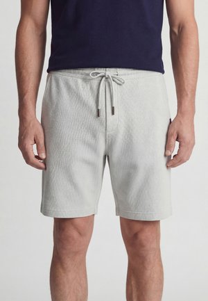 Man draagt lichtgrijze shorts met trekkoord en een donker marineblauw T-shirt, staat met ontspannen armen langs het lichaam tegen een effen achtergrond.