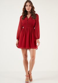 Rode chiffon jurk met een aansluitende lijfje, ronde halslijn en lange mouwen. Gerimpelde taille en uitlopende rok. Model draagt meerkleurige sandalen.