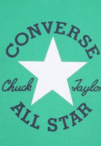 Vihreä t-paita, jossa on suuri valkoinen tähti ja musta teksti: "CONVERSE," "Chuck Taylor," ja "ALL STAR," tähden ympärille keskitettynä.