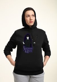 Persona che indossa un felpa nera con cappuccio alzato, mani in tasca, con una grafica che raffigura una figura che tiene un corvo e il testo "Nightshades & Ravens."