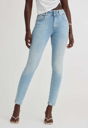 Person trägt hellblaue Skinny-Jeans und weiße Riemchensandalen, steht vor einem einfarbigen hellen Hintergrund.