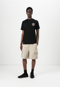 Svart bomull t-skjorte med rund logo, kombinert med beige cargo-shorts med sidelommer. Svart joggesko og ankelstrømper fullfører antrekket.