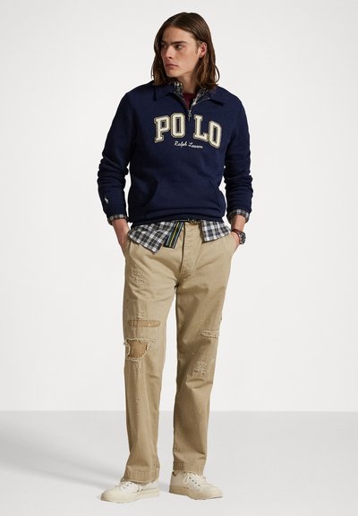 Mornarji pulover z logotipom "POLO Ralph Lauren", zložen čez karo srajco. Bež raztrgane hlače in bele superge dopolnjujejo obleko.