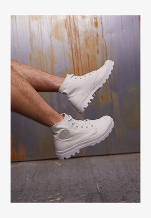 Weiße Canvas-Hochschaftstiefel mit grauer Gummisohle, Schnürung an der Vorderseite und rundem Logo-Patch an der Seite; strukturiertes Gewebe, leichtes Design.