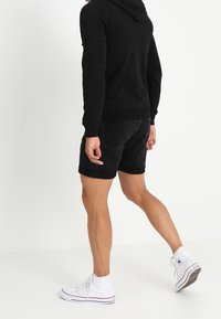 Sudadera negra con cordón, pantalones cortos de mezclilla negros y zapatillas altas blancas. El conjunto presenta un ajuste informal y relajado.
