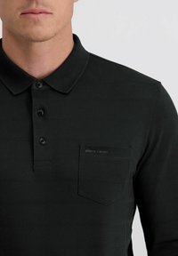 Schwarzes Poloshirt aus strukturiertem Stoff, mit einer Drei-Knopf-Leiste und einer Brusttasche, die ein dezentes Logo aufweist. Kurze Ärmel, klassisches Kragen-Design.