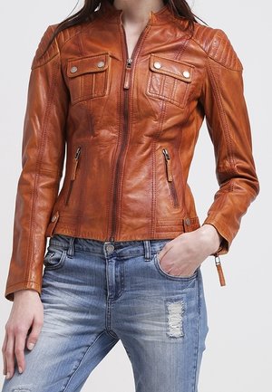 Vrouw draagt een nauwsluitend bruin leren jack met ritszakken en gewatteerde schouderdetails, gecombineerd met versleten blauwe spijkerbroek.