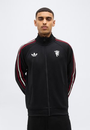 Mladý muž s krátkymi vlasmi, oblečený v čiernej bunde na zips s červenými a bielymi pruhmi na rukávoch a dvoma bielymi logami na hrudi.