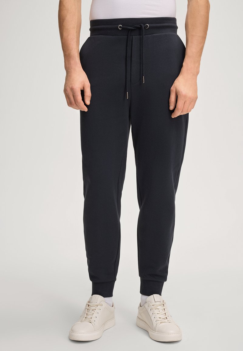 Navy Joggerhosen aus weichem Stoff, mit einem Kordelzugbund, seitlichen Taschen und gerippten Bündchen. Kombiniert mit cremefarbenen Sneakern.