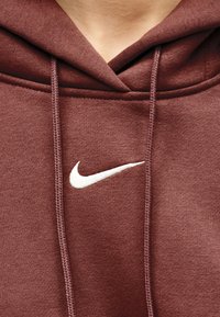 Κοντινό πλάνο ενός ατόμου που φοράει μια μαρόν μπλούζα με κουκούλα Nike με λευκό λογότυπο Swoosh και κορδόνια, εμφανίζοντας τον λαιμό και την ανώτερη περιοχή του στήθους.