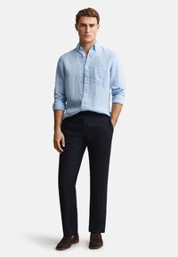 Jeune homme portant une chemise en lin bleu clair boutonnée, un pantalon foncé et des mocassins en daim marron, debout avec une main dans la poche.