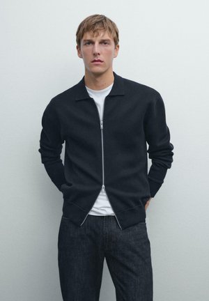 Un jeune homme portant une veste zippée marine sur une chemise blanche et un pantalon en jean foncé, se tenant devant un fond gris clair uni.
