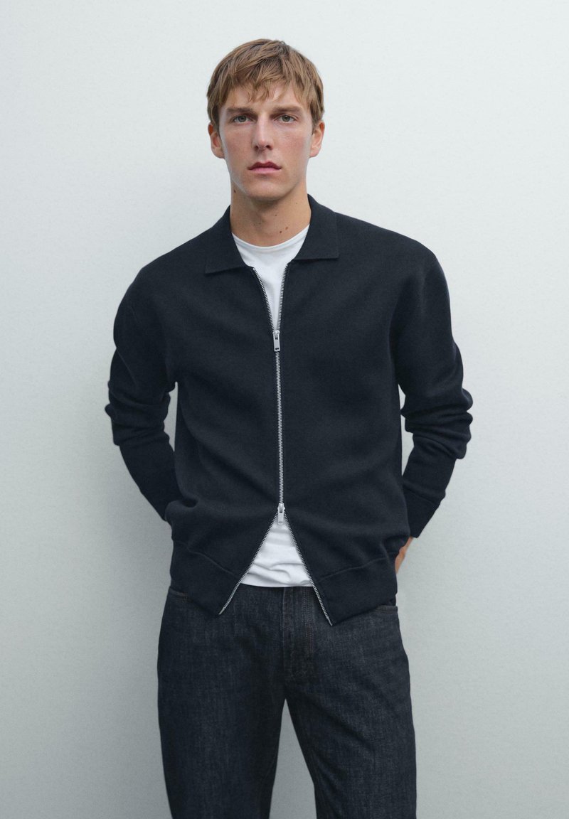 Junger Mann in einer marineblauen Zip-Jacke über einem weißen Hemd und dunklen Jeans, der vor einem schlichten hellgrauen Hintergrund steht.
