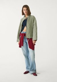Gestepptes grünes Jacket mit Rundhalsausschnitt, kombiniert mit einem marineblauen Crop-Top und lockeren hellblauen Jeans, mit einem kastanienbraunen Pullover um die Taille gebunden.