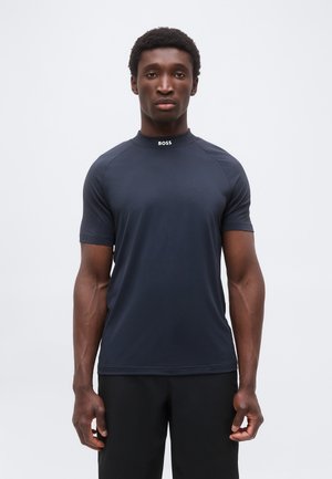 Homme debout, face à nous, portant une chemise ajustée bleu marine foncé à manches courtes avec le logo "BOSS" sur le col, sur un fond uni.