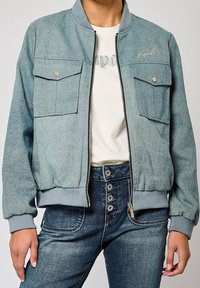Denim bomber jacket in faded blue with a ribbed collar, two front pockets, and zip closure. Worn over a white graphic t-shirt.