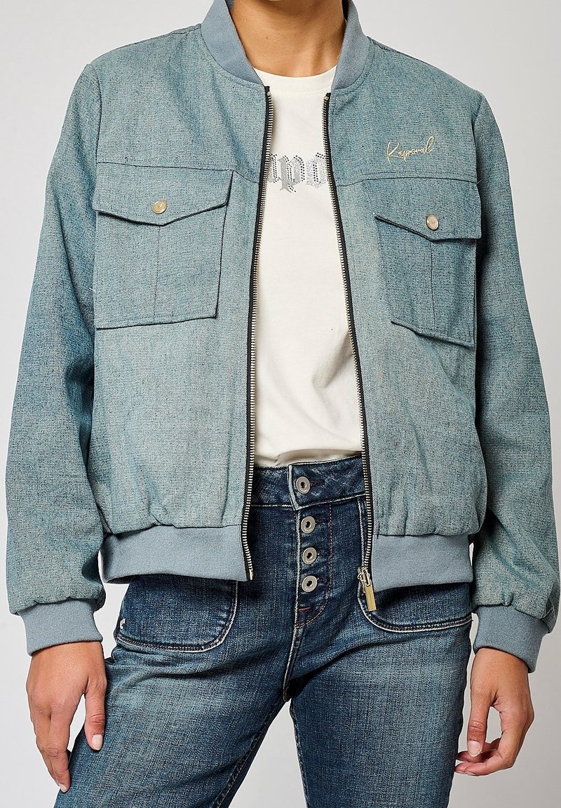 Denim bomber jacket in faded blue with a ribbed collar, two front pockets, and zip closure. Worn over a white graphic t-shirt.