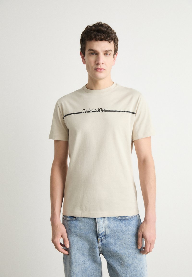 Beige bomull T-skjorte med en svart horisontal linje og "Calvin Klein"-logo. Korte ermer og rund hals, vist med lyseblå jeans.
