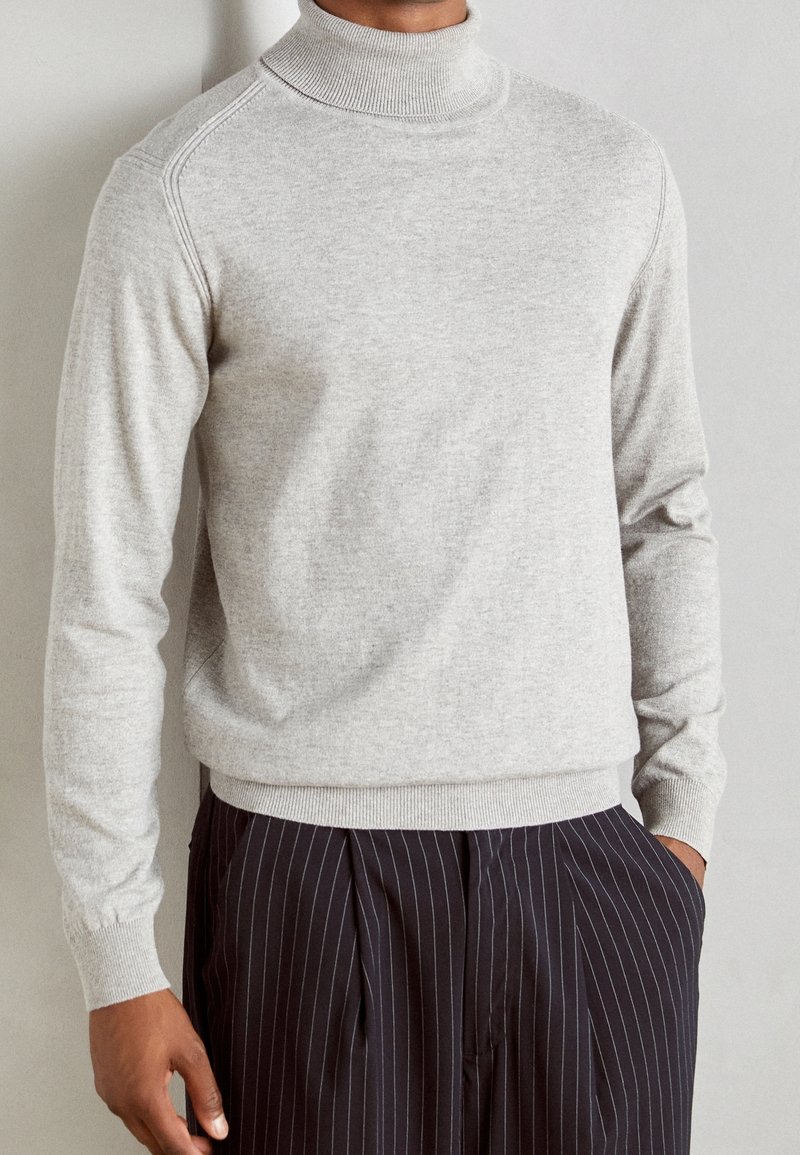 Lysegrå rullekravesweater med ribkant ved hals og ærmer og en glat tekstur. Lange ærmer og lige skæring, båret med mørke bukser med nålestriber.