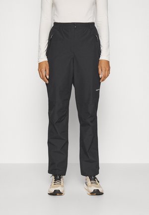 Persona vistiendo pantalones de exterior negros con cremallera y zapatos de senderismo beige, de pie frente a un fondo blanco liso.