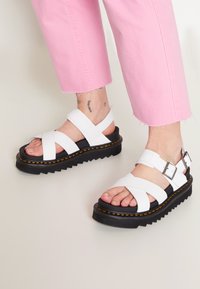 Weiße Plattform-Sandalen mit Kreuzriemen, verstellbaren Schnallen und einer strukturierten schwarzen Sohle mit gelben Nähten.