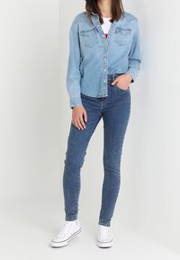 Camisa de mezclilla azul claro con botones en el frente y dos bolsillos en el pecho, combinada con jeans skinny azul oscuro y zapatillas blancas.