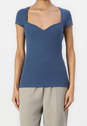 Vrouw draagt een aansluitende, korte mouw, geribd blauw topje met een sweetheart halslijn en losse beige broek.