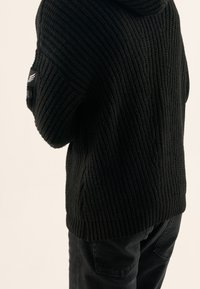 Svart stickad tröja med lös passform, med ribbad textur och huva. Inkluderar ett märke på vänster ärm. Bärs med svarta byxor.