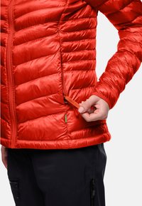 Giacca isolante rossa con texture trapuntata, dotata di tasca con zip e di un'etichetta arancione. Il tessuto ha una finitura lucida, offrendo calore.