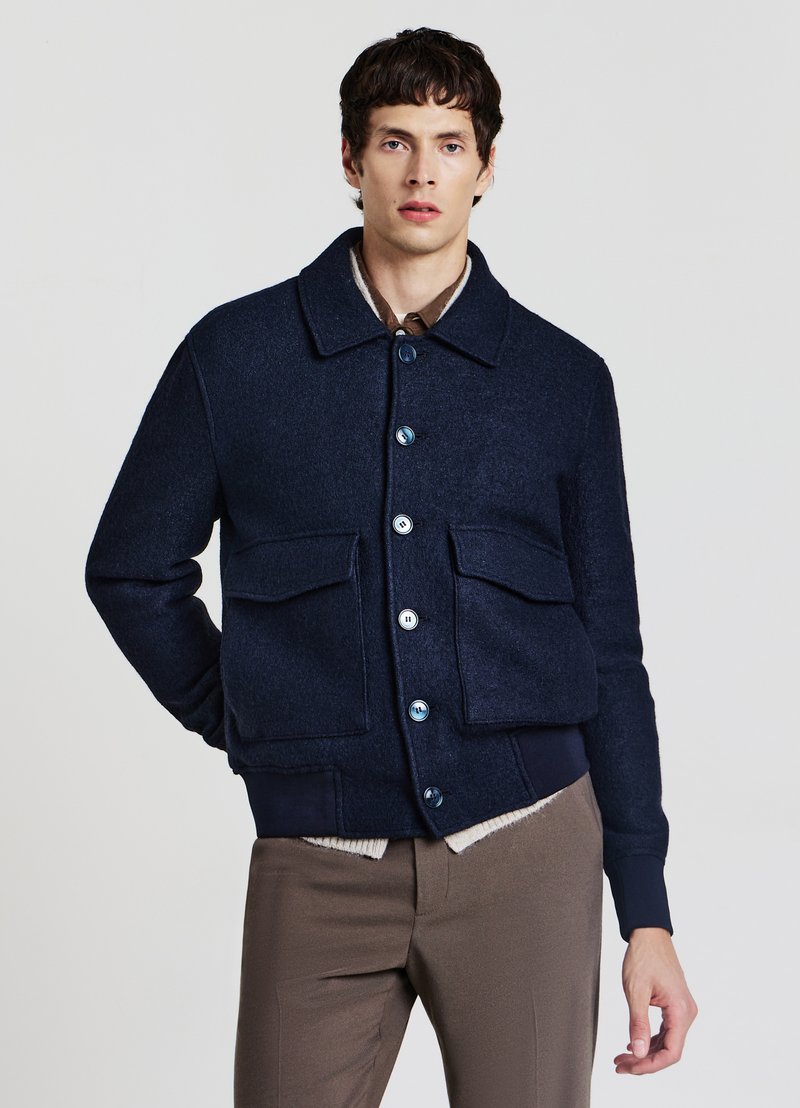 Giacca in misto lana blu navy con chiusura frontale a bottoni, grandi tasche frontali e polsini a coste. Presenta un colletto classico e una texture liscia.