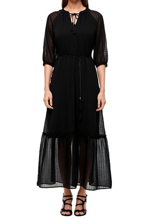 Femme portant une robe midi noire avec des manches transparentes et un voile sur la jupe, un col noué et une ceinture fine, associée à des talons noirs à brides.