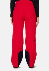 Pantalon de ski rouge avec une texture lisse, featuring des accents noirs au niveau des poignets et un patch logo à l'arrière. Conçu pour une utilisation active.